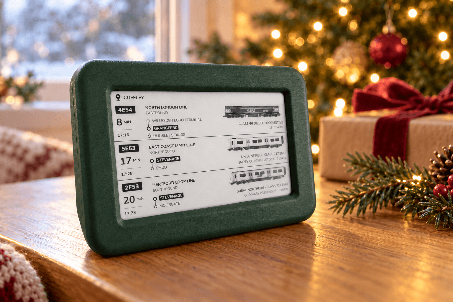 A green Lineside Ink display showing three trains at Cuffley on a wooden table beside a wrapped gift and a Christmas tree with glowing lights.