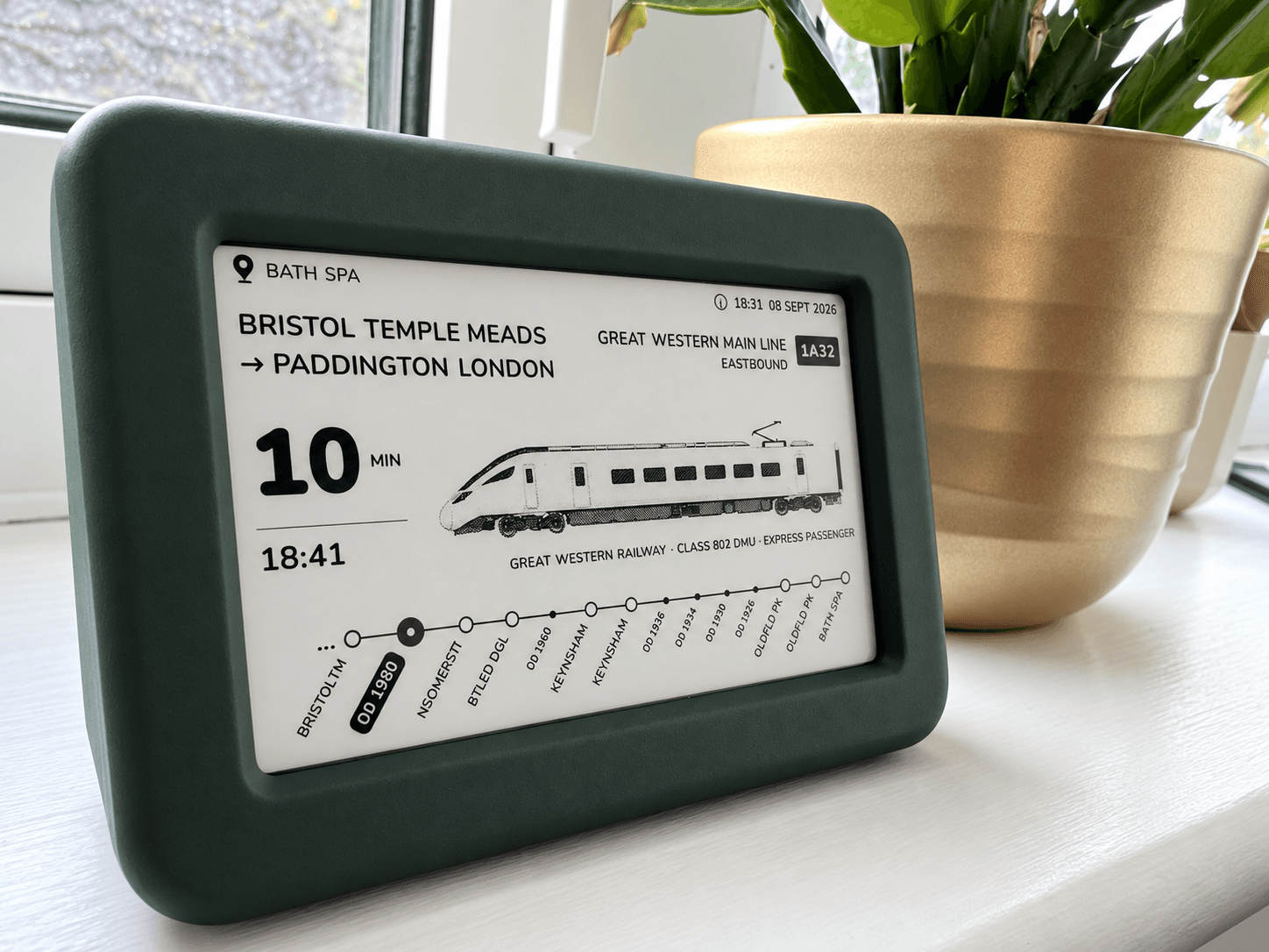 The green Lineside Ink display on a windowsill beside a houseplant, showing a Great Western service from Bristol Temple Meads to London Paddington approaching Bath Spa.