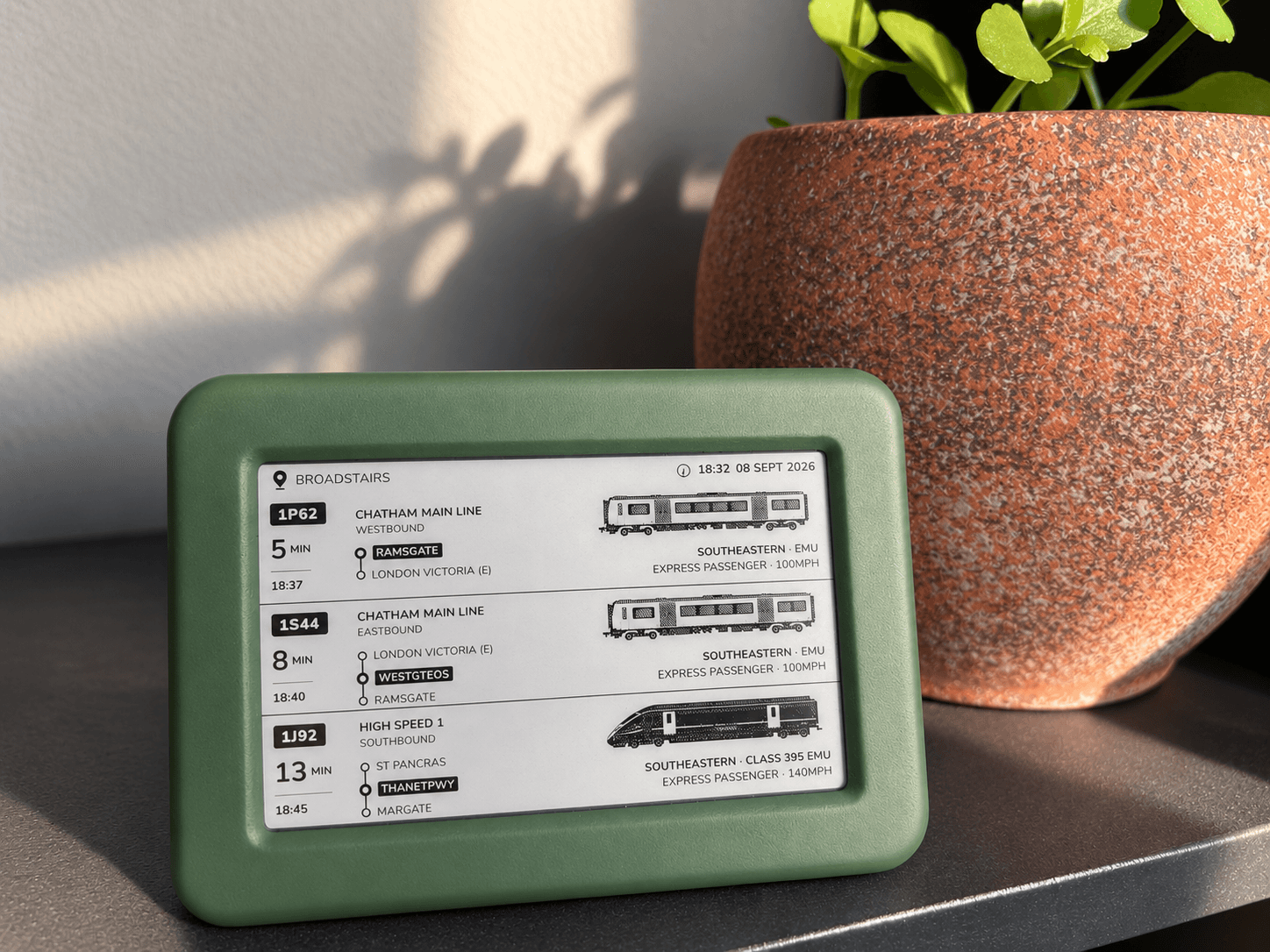 The green Lineside Ink display on a shelf beside a terracotta plant pot in dappled sunlight, showing approaching Southeastern services at Broadstairs.