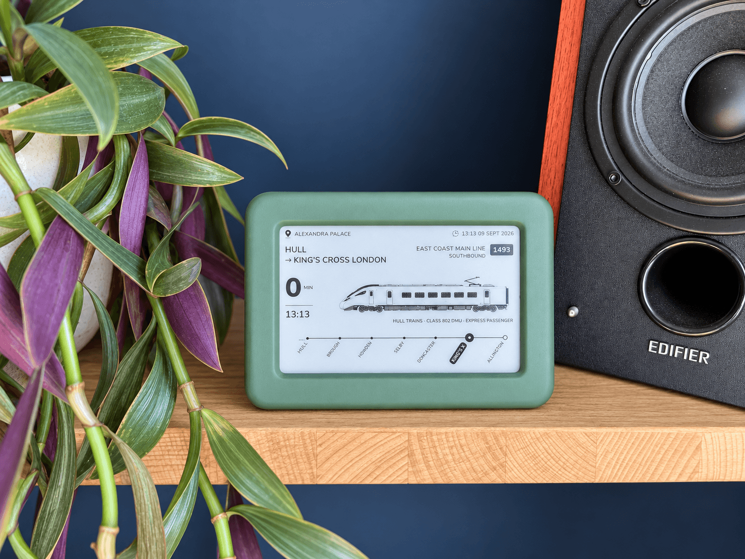 The Lineside Ink display in its deep green enclosure on a wooden shelf beside a hi-fi speaker and a trailing plant, its e-paper screen showing a Hull Trains service from Hull to King’s Cross on the East Coast Main Line.