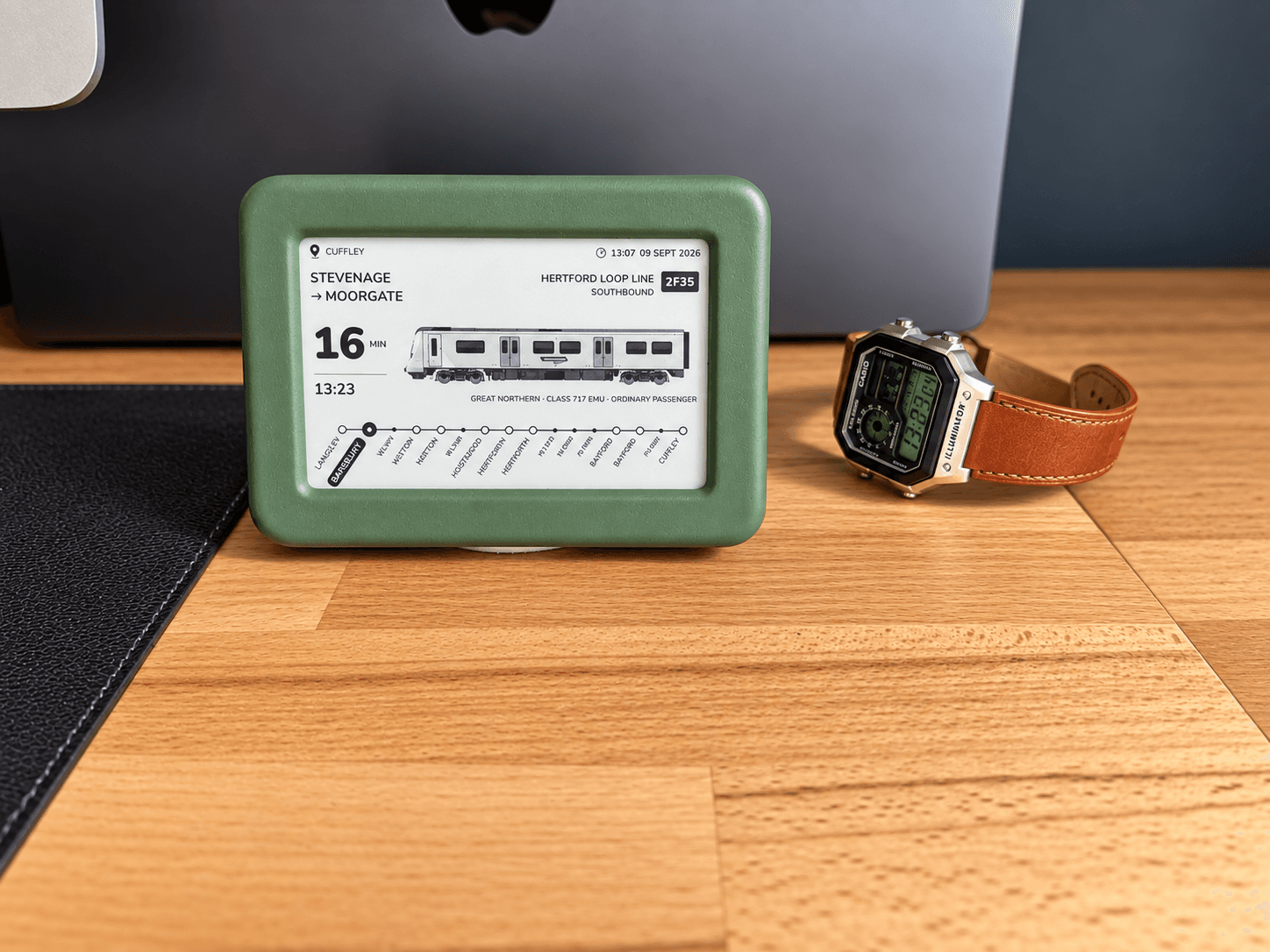 The green Lineside Ink display on a wooden desk beside a wristwatch and a laptop, its screen showing a Great Northern service from Stevenage to Moorgate on the Hertford Loop Line.