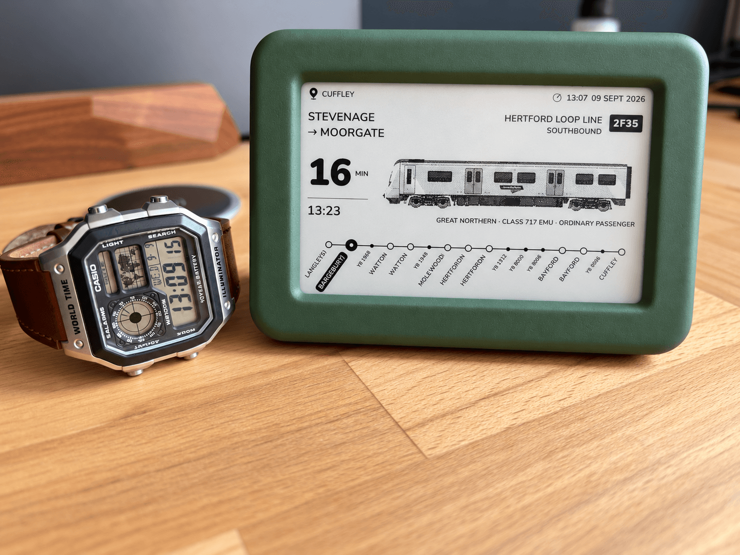 The green Lineside Ink display on a wooden desk beside a wristwatch, its screen showing a Great Northern service approaching Cuffley on the Hertford Loop Line.