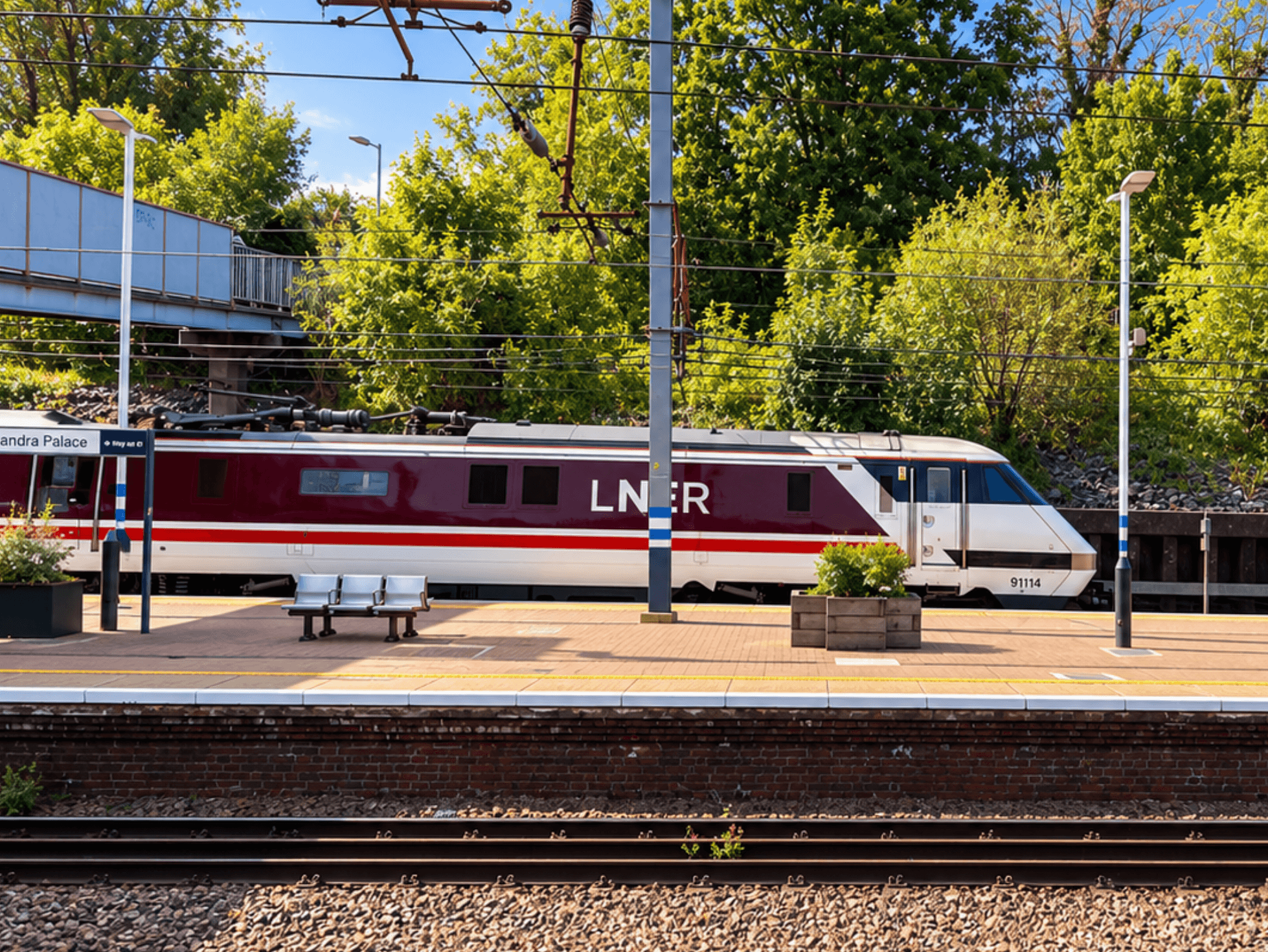 An LNER locomotive beside the platforms at Alexandra Palace station, beneath overhead wires and sunlit trees.