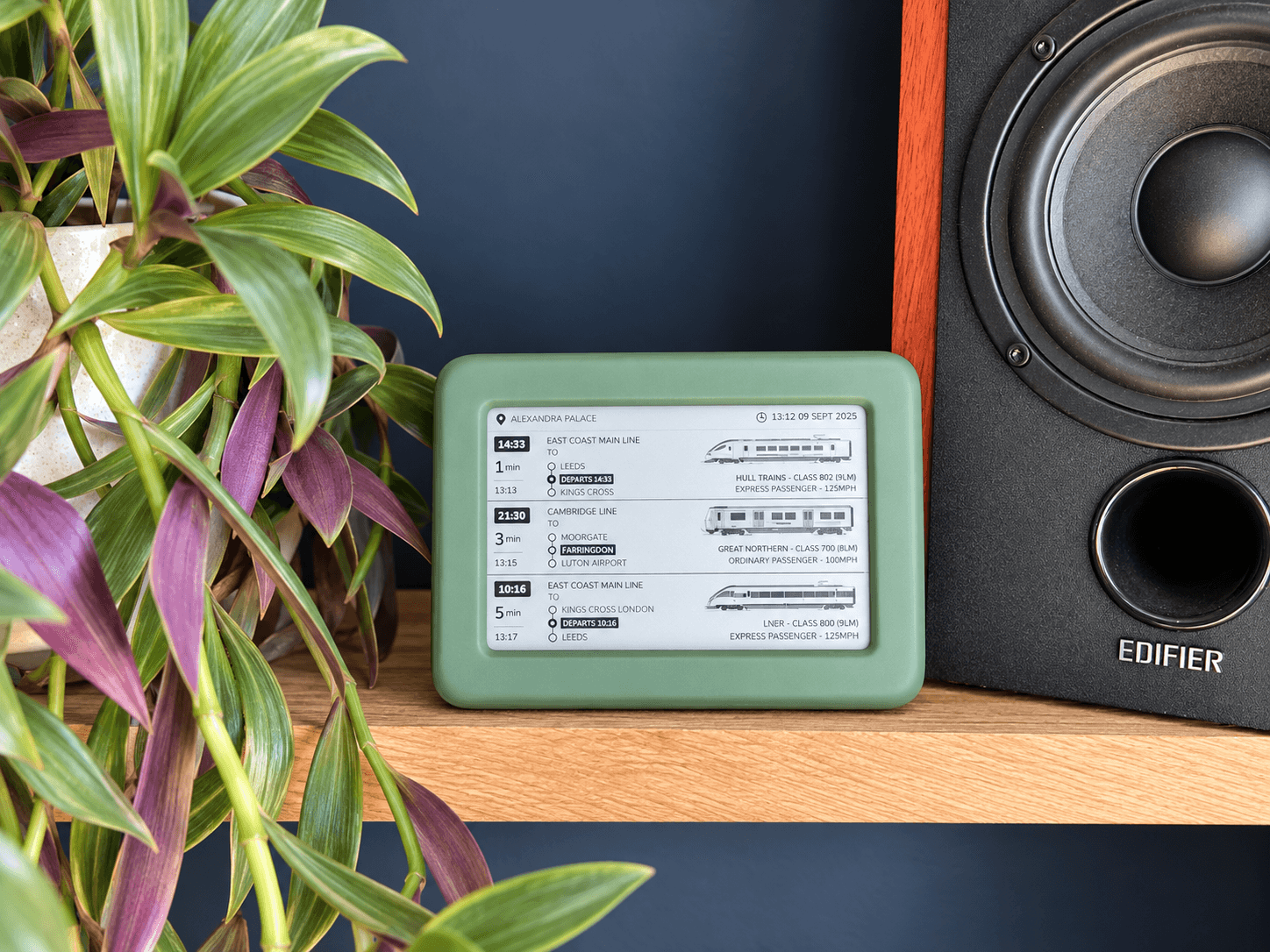 The green Lineside Ink display on a wooden shelf beside a houseplant and hi-fi speaker, its e-paper screen listing three approaching services at Alexandra Palace.