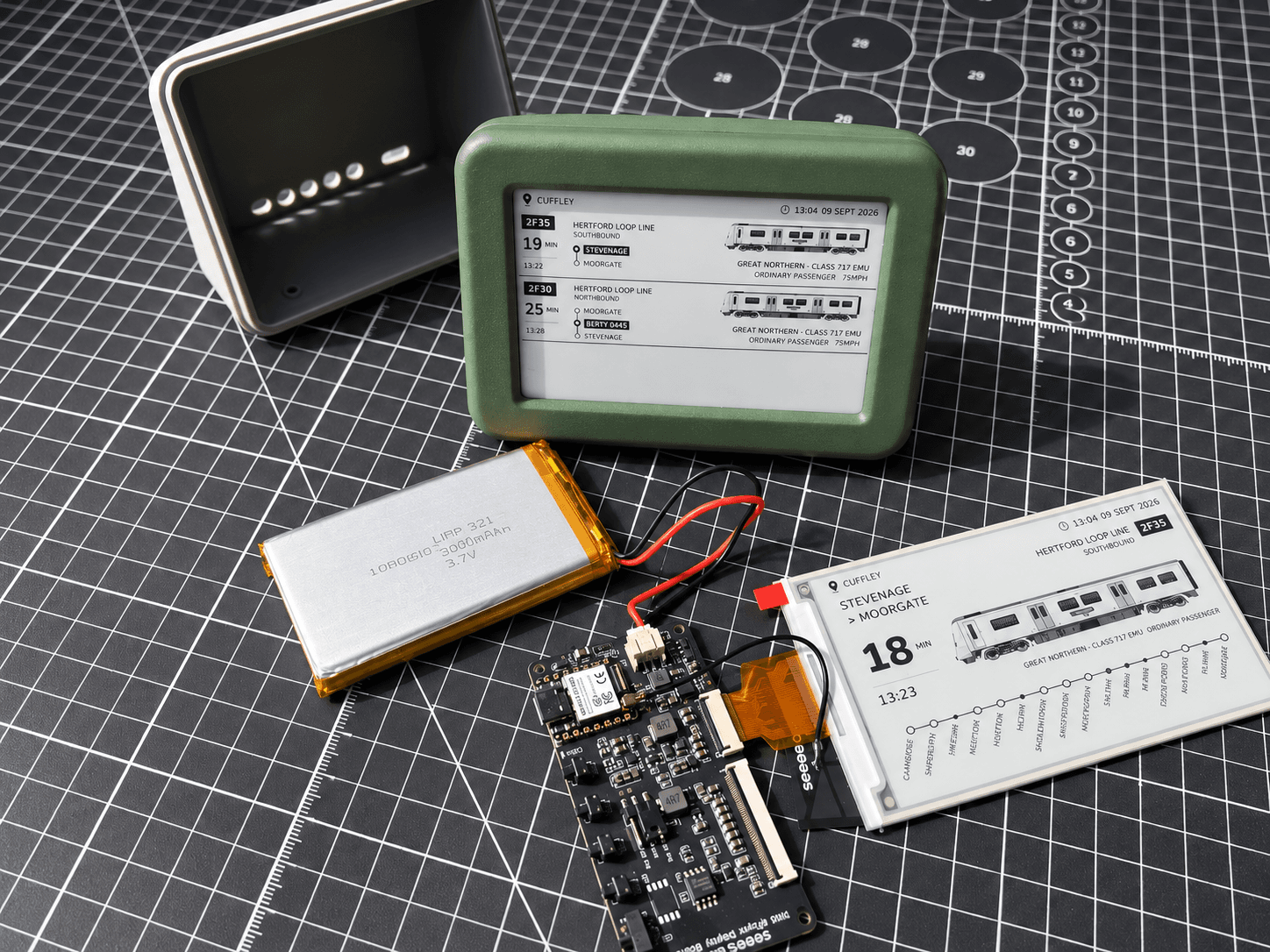 The parts of a Lineside Ink laid out on a cutting mat during assembly — the green 3D-printed enclosure, its rechargeable battery, the controller board and a bare e-paper panel showing a Hertford Loop Line service.