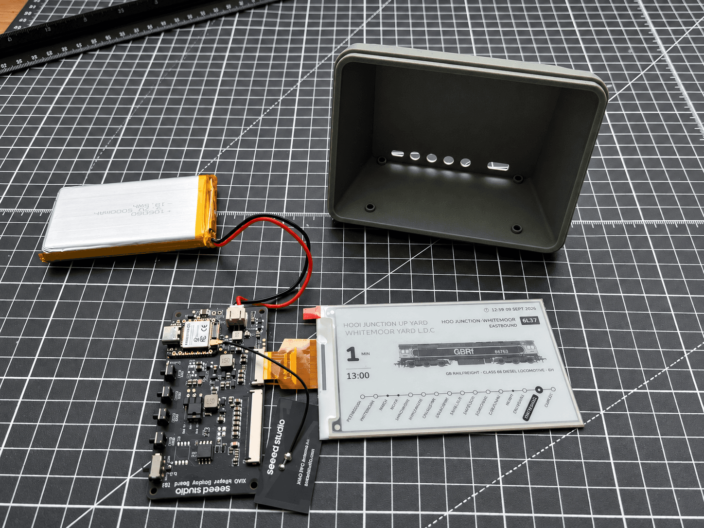 A Lineside Ink part-way through assembly on a cutting mat — the controller board and battery wired to a bare e-paper panel showing a GB Railfreight Class 66 approaching Whitemoor Yard, with the empty green enclosure behind.