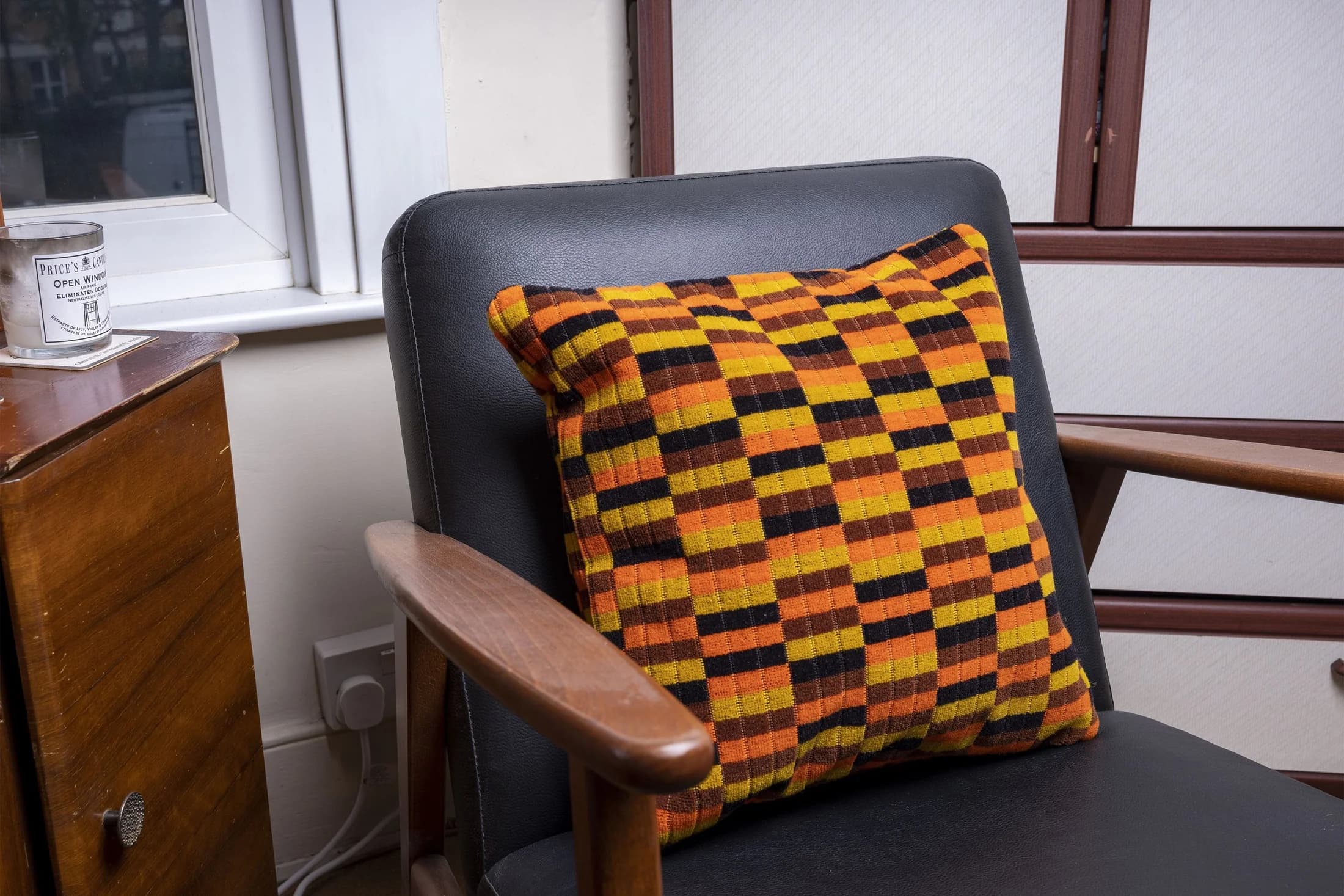 An orange, yellow, brown and black patterned moquette cushion on a dark armchair with wooden arms.